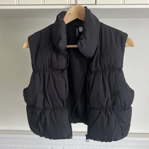 Svart dunväst från H&M - Säljer en svart dunväst från H&M med hög krage och dragkedja framtill. Västen har en puffig, quiltad design och två fickor på sidorna. Perfekt att slänga över en hoodie eller tröja för extra värme. Pris går att diskutera