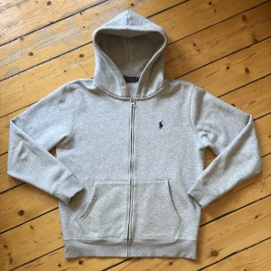 Grå hoodie från Polo Ralph Lauren - Säljer en klassisk grå hoodie från Polo Ralph Lauren. Tröjan har två fickor framtill och den ikoniska broderade loggan på bröstet. Perfekt för en avslappnad stil. Inte använd alls mycket ksk 3 gånger