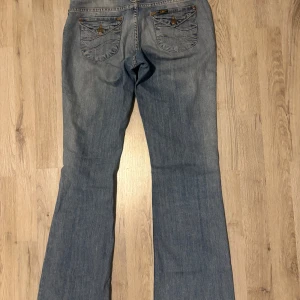 Blå bootcut jeans från Lee - Säljer ett par klassiska blå bootcut jeans från Lee med snygga detaljer på bakfickorna och normal passform. Jeansen har låg midja och är tillverkade i ett mjukt denimtyg. Perfekta för dig som gillar retrostil och vill ha ett par bekväma jeans.
