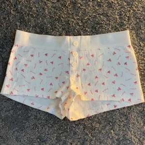 Vita shorts med rosetter och godiskäppar från Gina Tricot - Säljer ett par vita shorts från Gina Tricot med sött rosa mönster av rosetter och godiskäppar. Shortsen har bred resår i midjan och knappar framtill. Perfekta som mysplagg hemma. Har aldrig använt och ser helt nya ut. Skriv om du har några frågor!💗