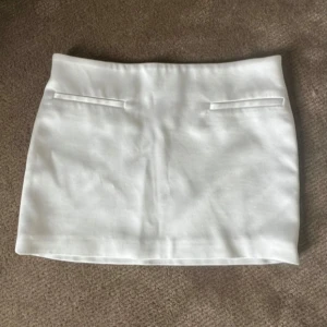 Vit kortkjol med fickdetaljer - Säljer en stilren vit kortkjol med två dekorativa fickor framtill. Kjolen har en rak passform och är perfekt för dig som gillar en enkel och clean look. Passar till många olika outfits och är lätt att matcha.