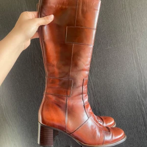 Läder höga stövlar med klack  - Säljer ett par cognac bruna höga stövlar i skinn med markerade sömmar och blockklack. Stövlarna har rund tå och dragkedja på insidan för enkel påtagning. Perfekta till hösten eller vintern till ett par jeans. De har ett rutigt mönster vilket gör dem lite unika, älskar dem men rensa lite skor hemma. Kommer hålla hålla hålla! 