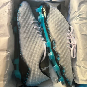 Nike hypervenom - Snygga fotbollsskor i vitt med turkosa och svarta detaljer. Skorna har en texturerad ovandel och turkos sula med dobbar för bra grepp på planen. Klassisk snörning och modern design som sticker ut på matchen.