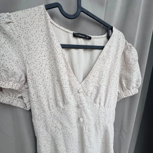Ljusprickig långklänning från Stinna - Supersöt ljus långklänning från Stinna med små svarta prickar, v-ringning och puffärmar. Klänningen har knappar framtill och resår vid ärmslut. Perfekt för sommarens alla tillfällen!
