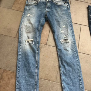 Blå slitna jeans från Replay Grover - Säljer ett par blå Replay Grover jeans med raka ben och slitna detaljer framtill. Jeansen har klassisk femficksdesign, dragkedjegylf och tydliga slitningar för en tuff look. Perfekta för dig som gillar en avslappnad stil.