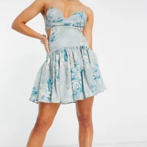 Blommig ljusblå kortklänning med smala axelband - Supersöt kortklänning i ljusblått med blommigt mönster och smala axelband. Klänningen har en markerad midja, lätt utställd kjol och cut-out detaljer vid sidorna. Perfekt för varma dagar och somriga tillfällen.