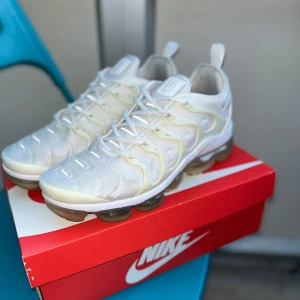 Nike Air Vapormax Plus vita sneakers - Nike Air Vapormax Plus sneakers i vitt med transparent sula och vita detaljer. Skorna har snörning och en unik, vågig design på ovandelen. Skorna kommer i orginalkartong. Perfekta för dig som gillar en modern och sportig look.