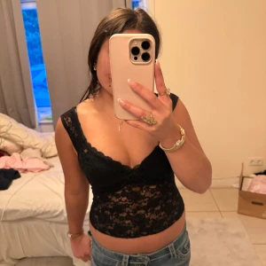 Svart spetsig croppad linnetopp - Säljer en svart croppad linnetopp i spets med v-ringning. Toppen är ärmlös och har ett transparent blommigt spetsmönster. Perfekt för att skapa en trendig och feminin look.