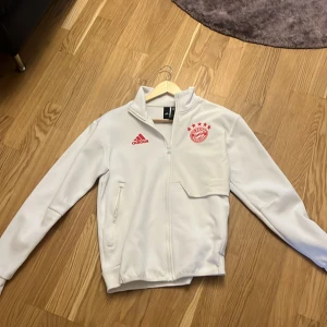Vit Adidas FC Bayern München jacka - Säljer en vit träningsjacka från Adidas med FC Bayern München-logga på bröstet. Jackan har dragkedja, hög krage och två fickor framtill. Perfekt för fotbollsträning eller som supporterplagg. Adidas-logga i rött på bröstet.