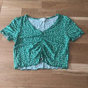 Grön blommig croppad topp - Grön croppad topp med små vita blommor, v-ringning och rynkade detaljer framtill. Korta ärmar med vågig kant. Från Gina i storlek xs