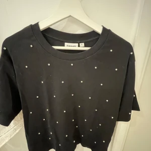 Svart t-shirt med pärlor från Kappahl - Svart t-shirt från Kappahl dekorerad med små pärlor framtill. T-shirten har rund halsringning och korta ärmar. Perfekt för dig som vill ha en enkel men ändå lite extra topp.