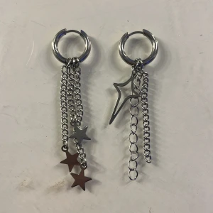 Silverfärgade örhängen med stjärnhängen och kedjor - Örhängen i silverfärg med hängande kedjor och stjärndetaljer. Ena örhänget har tre små stjärnor och det andra har en större stjärna och kedjor i olika längder. Perfekt för dig som gillar unika accessoarer.