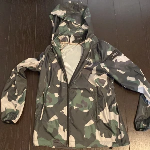 Camouflage vindjacka från Orkan - Säljer en snygg vindjacka från Orkan i grön, svart, vit och grå camouflage. Jackan har huva, dragkedja framtill och elastiska muddar vid ärmsluten. Perfekt för blåsiga dagar och streetwear-stil. Ny pris 1499!