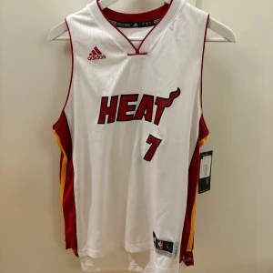Miami Heat baskettröja Dragic 7 - Säljer en vit Miami Heat baskettröja från Adidas med röd och gul detalj på sidorna. Nummer 7 och namnet Dragic på ryggen. Klassisk NBA-design med HEAT-logga framtill och ärmlös modell.