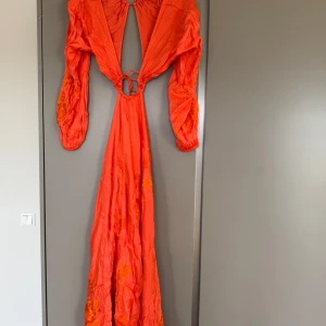 Orange långklänning med broderade detaljer - Säljer en orange långklänning med broderade blommor och långa ärmar. Klänningen har djup v-ringning, öppen rygg, midja och knytning framtill med en dekorativ ring. Perfekt för dig som vill sticka ut med färg och unika detaljer.