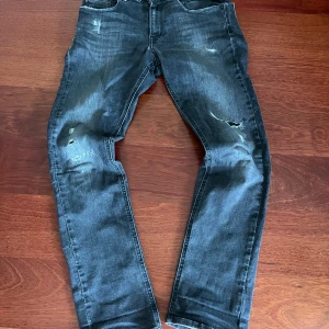Replay svarta jeans med slitningar 150 cm - Säljer ett par svarta Replay jeans med slitna detaljer på benen och klassisk femficksdesign. Jeansen har normal passform och är tillverkade i denim med Replay-logga vid fickan och bak på linningen.
