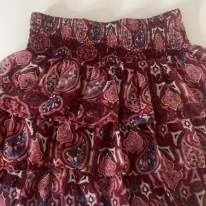 Mönstrad volangkjol från Lindex - Säljer en färgglad kjol från Lindex med paisleyinspirerat mönster i vinrött, rosa, blått och orange. Kjolen har bred resår i midjan och flera volanger för en luftig känsla. 