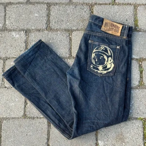 Mörkblå jeans från Billionaire Boys Club med astronauttryck - Säljer ett par mörkblå jeans från Billionaire Boys Club med astronauttryck på bakfickan och snygga detaljer som broderad logga i midjan och på knappen. Klassisk femficksmodell med raka ben och läderpatch bak.