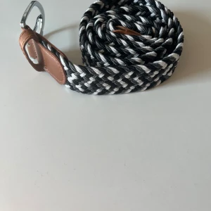 Svart och vit flätat bälte med läderdetaljer - Säljer ett snyggt flätat bälte. Bältet har ett silvrigt metallspänne och passar perfekt till jeans eller chinos. Aldrig använt, helt i nyskick.