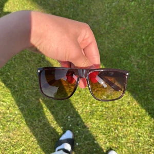 Bruna fyrkantiga solglasögon lika raybans - Snygga solglasögon med breda, bruna bågar och fyrkantig form. Glasen är mörka och ger ett coolt intryck. Perfekta för att lyfta din stil under soliga dagar.