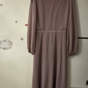 Långklänning med broderade blommor och v-ringning - Långklänning i dov lila nyans med v-ringad hals och långa ärmar. Klänningen har vackra broderade blommor i rosa, blått och beige framtill, vilket ger en romantisk känsla. Perfekt för dig som vill sticka ut med detaljer.