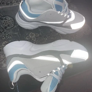 Grå och vita sneakers från Dior - Säljer ett par snygga Dior sneakers i vitt, grått och ljusblått. Skorna har en chunky sula, meshdetaljer och reflekterande partier. Perfekta för dig som vill sticka ut med stilrena och moderna sneakers. Helt nya ‼️med kvitto 