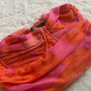 Blommig tubtopp från Zara - Säljer en färgstark tubtopp från Zara med blommigt mönster i orange och rosa nyanser. Toppen har rynkade detaljer framtill och är axelbandslös.