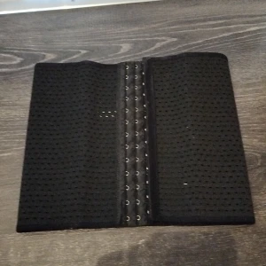 Svart korsett waisttrainer med hyskknäppning - En svart korsett med perforerat tyg och flera rader hyskor för justerbar passform. Perfekt för att forma midjan eller som accessoar till olika outfits. Storlek small 62-68 cm