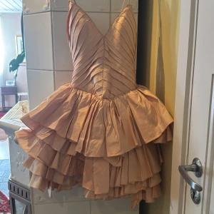 Champagnefärgad volangklänning med v-ringning - Säljer en champagnefärgad kortklänning med djup v-ringning och tunna axelband. Klänningen har flera lager volanger och en omlottliknande effekt framtill. Perfekt för dig som vill sticka ut med en unik och festlig look.