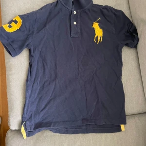 Mörkblå pikétröja från Polo Ralph Lauren - Säljer en mörkblå pikétröja från Polo Ralph Lauren med stor gul broderad logga på bröstet och siffran 3 på ärmen. Klassisk krage och knappar framtill. Perfekt för en sportig och stilren look. 