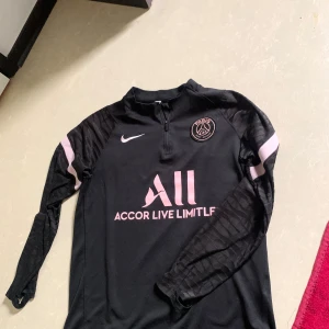 Svart långärmad Paris Saint-Germain fotbollströja från Nike - Säljer en svart långärmad PSG-tröja från Nike med rosa detaljer och klubbmärke på bröstet. Tröjan har dragkedja vid halsen och texten 'Accor Live Limitless' samt 'Visit Rwanda' på ryggen. Perfekt för dig som älskar fotboll och vill representera Paris Saint-Germain.