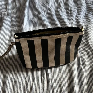 Svart och guld randig väska - Väska/necessär med svarta och guldiga ränder. Väskan har dragkedja upptill och ett praktiskt handledsband i guld. Ca 20cm lång & 11cm hög.