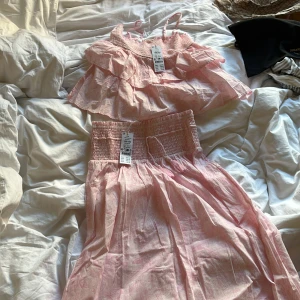 Ljusrosa tvådelad klänning med volanger - Säljer en söt tvådelad klänning i ljusrosa med smockdetaljer och volanger. Toppen har smala axelband och volang, kjolen är lång med smock i midjan. Perfekt för sommarens alla tillfällen!