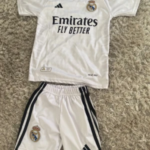 Real Madrid fotbollströja och shorts - Säljer en vit Real Madrid fotbollströja med svarta detaljer och tillhörande shorts. Tröjan har klubbmärke och sponsortryck framtill samt svarta ränder på axlarna. Shortsen har klubbmärke och svarta ränder längs sidorna. Perfekt för fotbollsträning eller match.