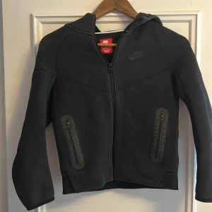 Svart zip hoodie från Nike - Säljer en svart zip hoodie från Nike med dragkedja framtill och två fickor med dragkedja. Jackan har huva och en stilren design med diskret Nike-logga på bröstet. Perfekt för en sportig och avslappnad look. Den är i barnstorlek M som är storlek 137-147 cm