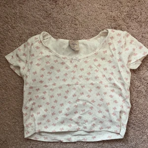 Vit croppad topp med små rosa blommor från Rosemunde - Supersöt vit croppad topp från Rosemunde med små rosa blommor och en liten rosett vid halsringningen. Toppen har korta ärmar och rund hals. Perfekt för en söt och romantisk look. Skulle säga storlek M-L