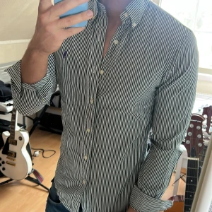 Grön- och vitrandig skjorta från Ralph Lauren - Säljer en slim fit skjorta från Ralph Lauren i grönt och vitt med klassiska ränder. Skjortan har lång ärm, knappar framtill och en broderad lila logga på bröstet. Perfekt för dig som gillar stilrena plagg med snygga detaljer.