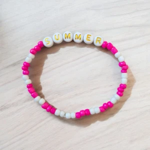 Färgglatt armband - Färgglatt armband med text – "SUMMER"  Ge din outfit en somrig och lekfull touch med detta handgjorda armband! Prydligt designat med vita och knallrosa pärlor samt bokstavspärlor som stavar ut ordet "SUMMER" i klara guldiga bokstäver. Det elastiska bandet gör det enkelt att ta på och av, samtidigt som det sitter bekvämt runt handleden.  Material: Plastpärlor  Färg: Vit, rosa och guld  Text: SUMMER  Storlek: One size, elastisk passform