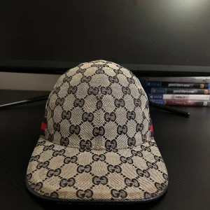 Gucci keps med GG-mönster och röd-blå rand - Snygg keps från Gucci med klassiskt GG-mönster i beige och svart. Kepsen har en röd och blå rand på sidan och justerbar rem baktill. Tillverkad i bomullsmaterial med svart insida. Perfekt för dig som vill sticka ut med en lyxig accessoar.