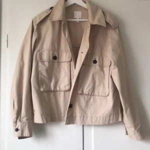 Beige jacka med knappar - Snygg beige vårjacka med stora fickor och knappar framtill. Jackan har en klassisk krage och långa ärmar. Perfekt för en stilren look. Köpt från H&M och är i strl S. Knappt använd