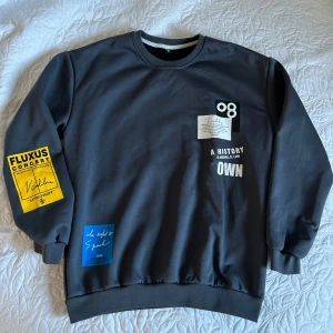 Grå sweatshirt med tryck  - Snygg grå sweatshirt med flera tryck och detaljer från AFYF. Tröjan har långa ärmar och en rund halsringning. Den har unika tryck i vitt, gult och blått med text och grafiska element. Perfekt för en cool och avslappnad stil.