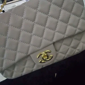 Beige quiltad axelväska - Snygg beige axelväska med quiltat mönster och gulddetaljer. Perfekt för att ge en elegant touch till din outfit. Väskan har en klassisk design med en vridbar låsning framtill.