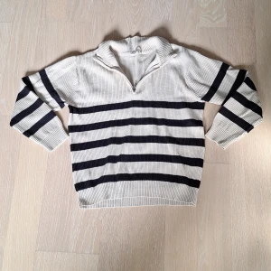 Randig stickad tröja med half-zip - Säljer en beige stickad tröja med svarta horisontella ränder. Tröjan har lång ärm, hög krage och kort dragkedja framtill. Perfekt för en avslappnad och stilren look.