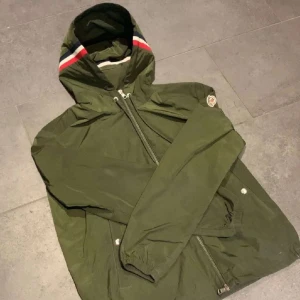 Grön vindjacka med huva från Moncler - Snygg grön vindjacka från Moncler med huva och dragkedja framtill. Jackan har vita och röda ränder på huvan och Moncler-logga på ärmen. Perfekt för blåsiga dagar och enkel att matcha med det mesta.