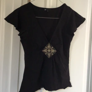 Svart kortärmad topp från att. - Snygg svart topp från att. med korta ärmar och v-ringning. Den har en dekorativ broderad detalj framtill i form av en fyrkant. Perfekt för en stilren look.