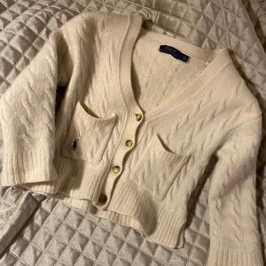 Vit kabelstickad kofta från Polo Ralph Lauren - Säljer en stilren vit kabelstickad kofta från Polo Ralph Lauren. Koftan har en v-ringad design med knappar framtill och två praktiska fickor. Den är tillverkad i ett mjukt kvalitativt material av ull och kashmir.