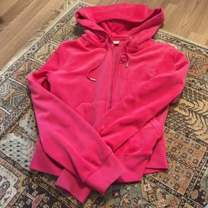 Rosa velourhoodie från Juicy Couture - Juicy Couture hoodie i starkt rosa velour med dragkedja och huva. Guldiga detaljer på snören och dragkedja, samt broderad JC-logga på bröstet. Perfekt för en sportig och trendig look. Äktaaa