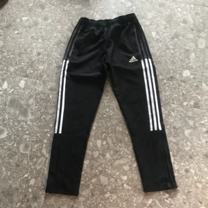 Svarta träningsbyxor från Adidas med vita ränder - Svarta träningsbyxor från Adidas med klassiska tre vita ränder längs sidorna och elastisk midja. Byxorna har AEROREADY-teknologi som transporterar bort fukt, perfekt för sport och aktivitet. Adidas-logga framtill.