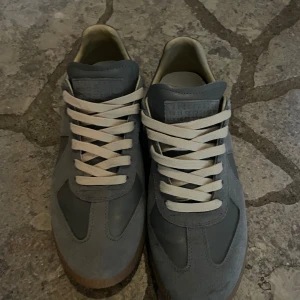 Grå sneakers från Maison Margiela  - Säljer ett par grå Maison Margiela sneakers med vita snören. Skorna har en klassisk design med paneler i mocka och skinn, samt rund tå. Perfekta för dig som gillar stilrena och tidlösa sneakers. Skorna är i storleken 43 men passar utmärkt till 42. OBS! Har inget kvitto tyvärr slängde det 