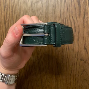 Mörkgrönt skärp från Gucci - Säljer ett mörkgrönt skärp från Gucci i skinn med präglad GG-logga runt om. Skärpet har en silverfärgad metallspänne och bred design. Perfekt för att lyfta din outfit med en exklusiv touch.